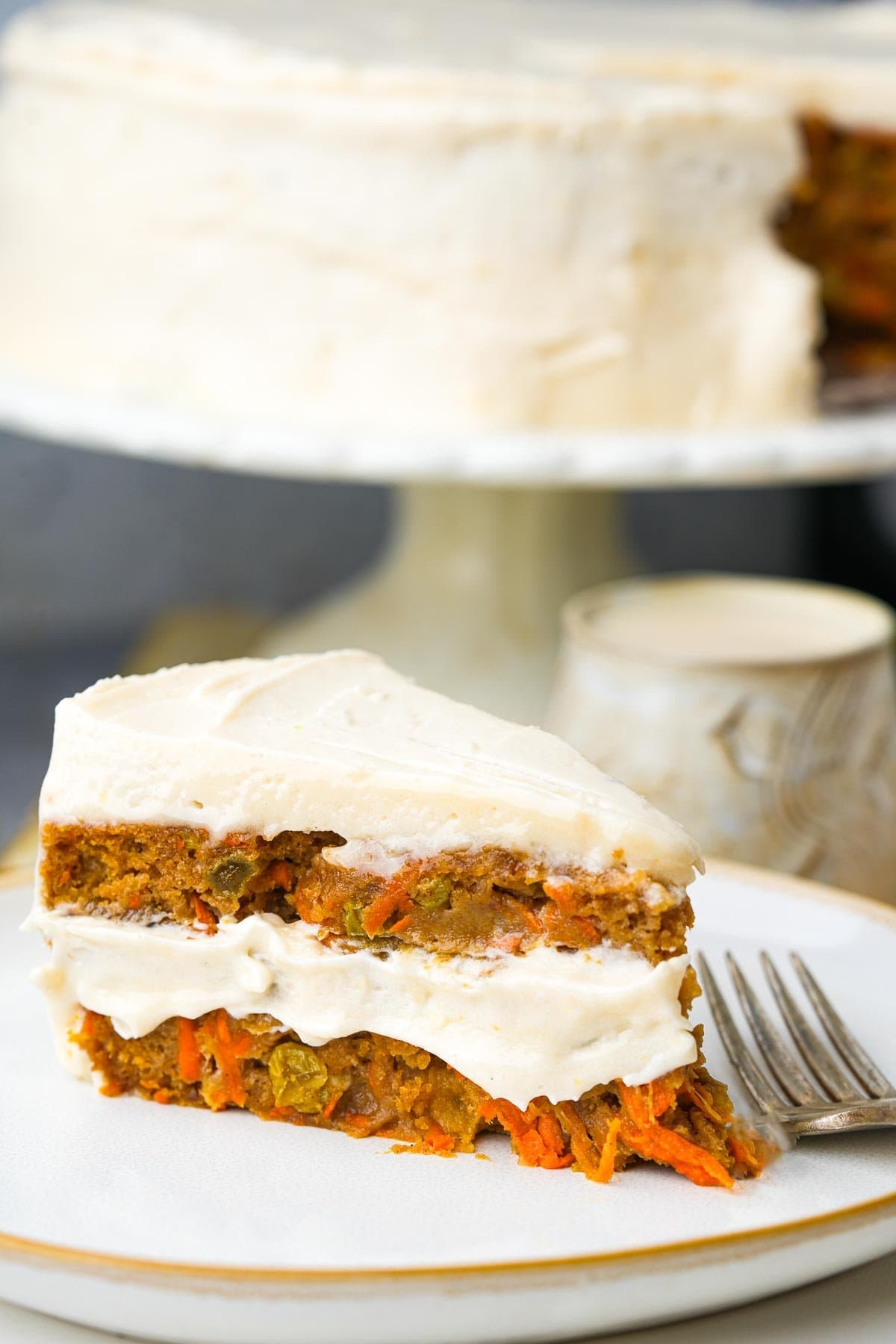 A slice of vegan carrot cake with cream cheese frosting on a white plate, with the whole cake visible in the background.