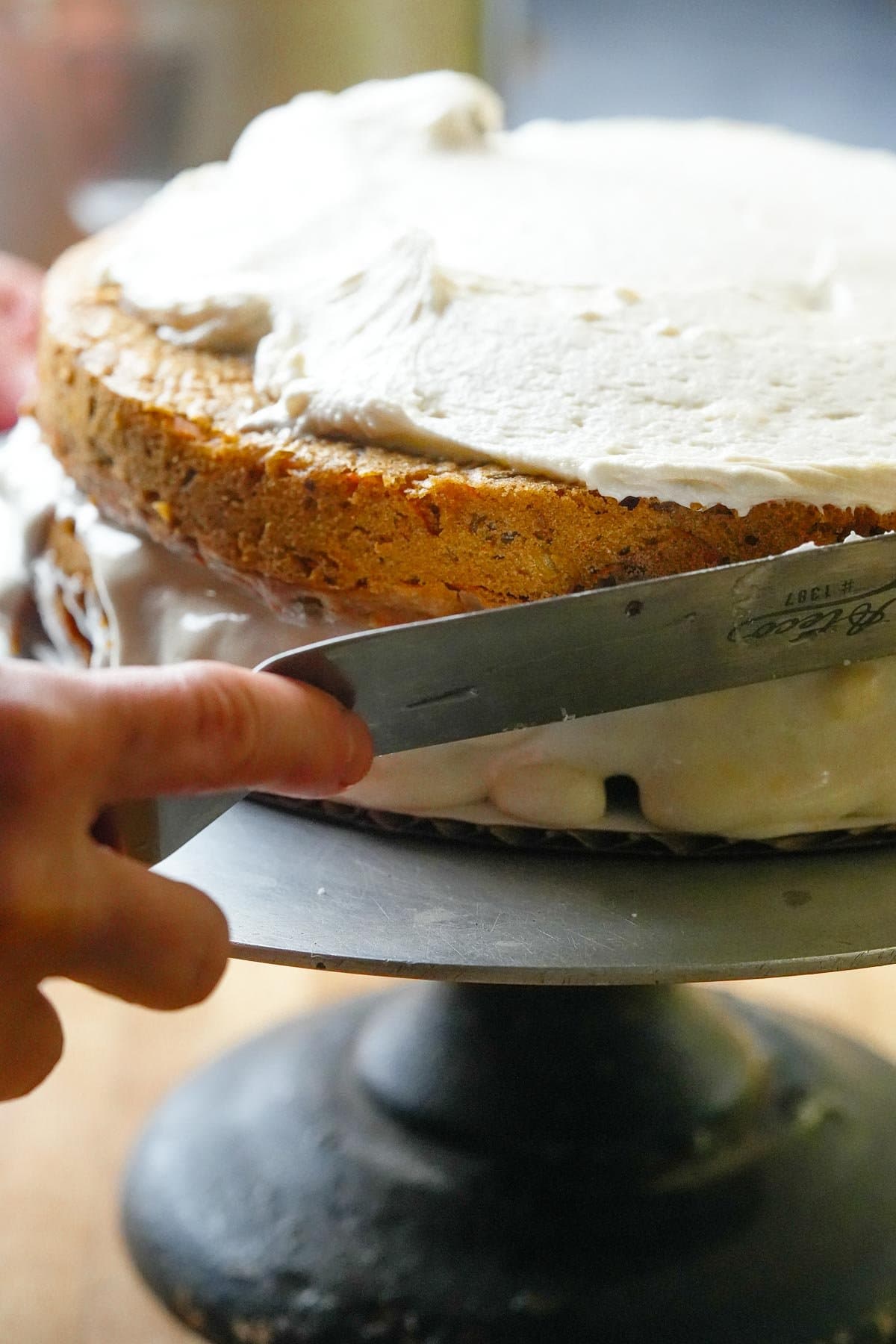 A person frosting a cake, holding a spatula with white icing, placed on a rotating cake stand.