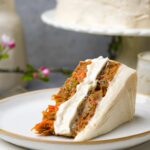 A slice of vegan carrot cake with cream cheese frosting on a white plate, garnished with edible flowers, beside a whole cake.