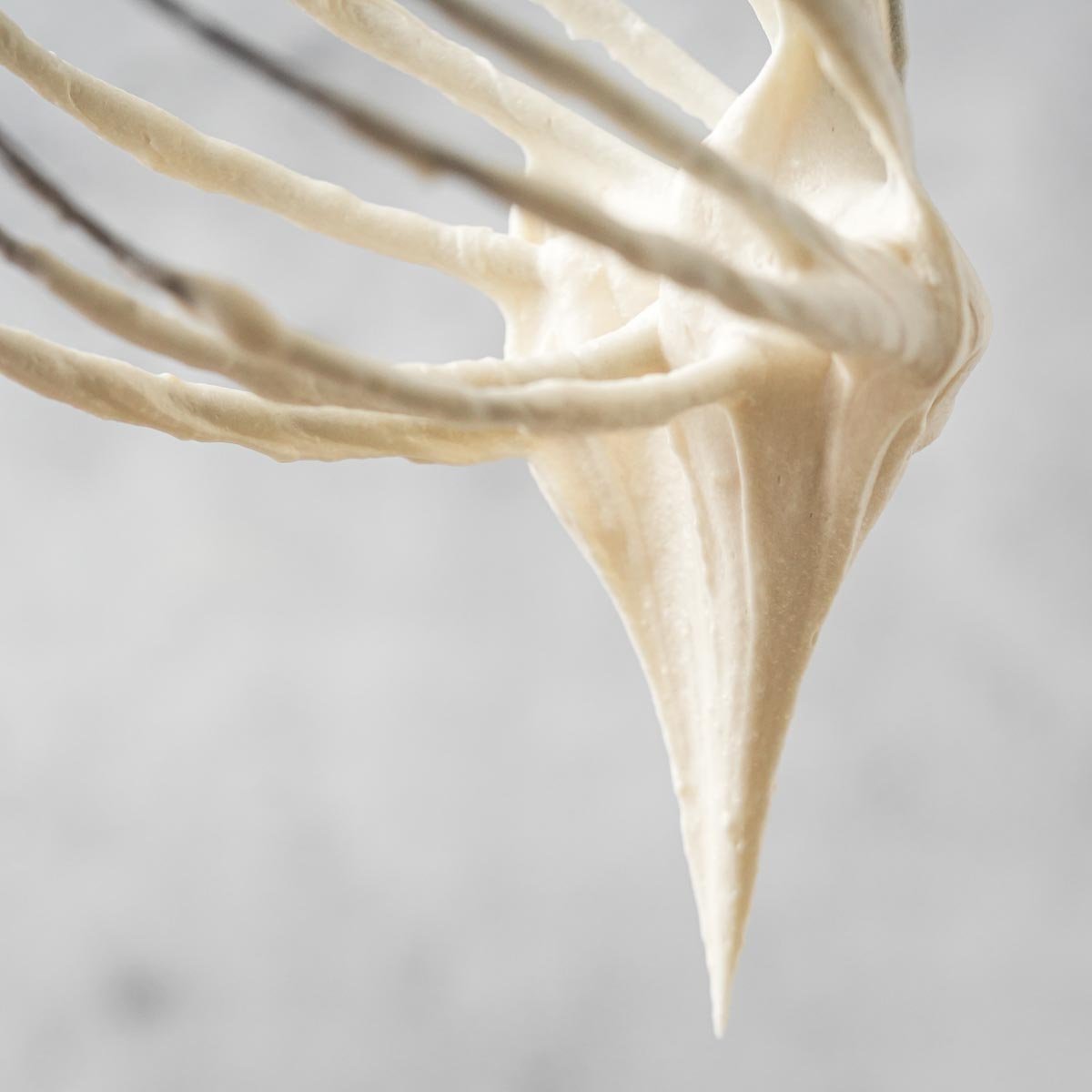 Whisk with Vegan cream cheese frosting dripping off, against a blurred light background.