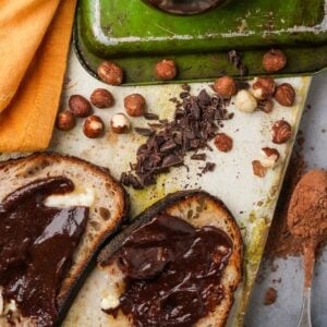 Slices of bread topped with chocolate spread, hazelnuts, and chopped chocolate on a kitchen counter.