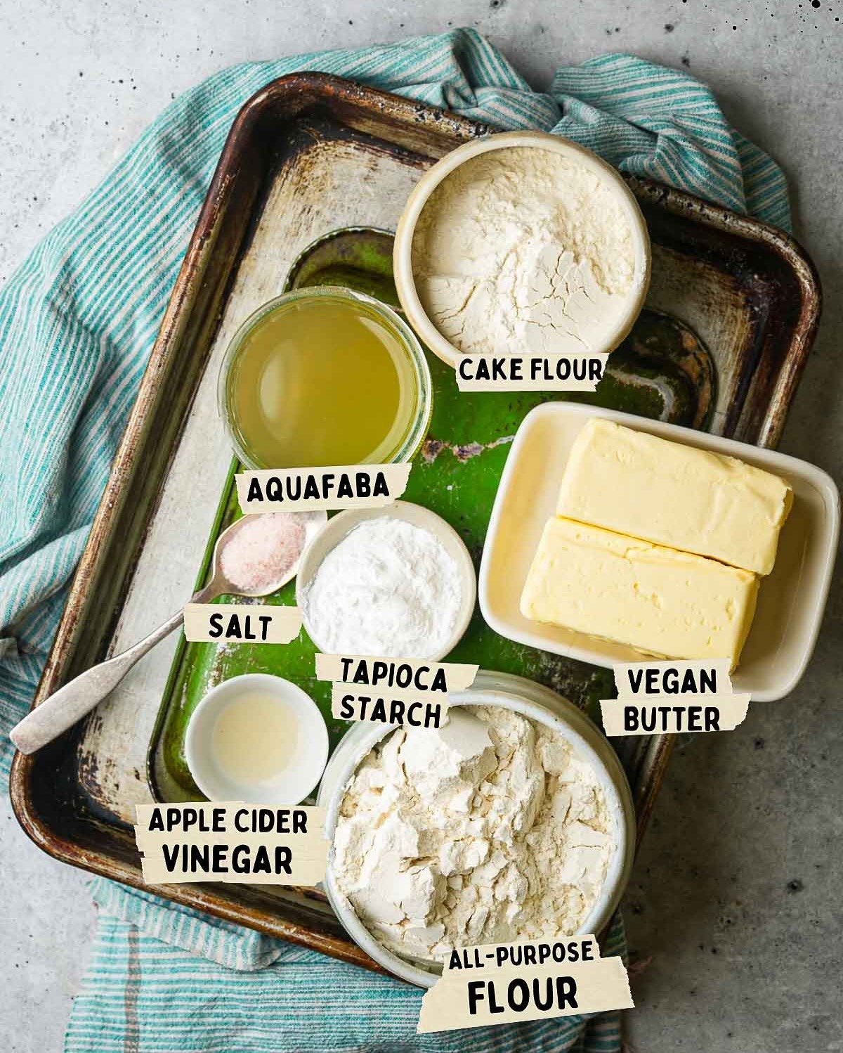 A baking tray with labeled ingredients for a vegan pie crust: cake flour, vegan butter, all-purpose flour, tapioca starch, aquafaba, salt, apple cider vinegar on a green towel background.