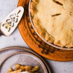 Freshly vegan baked apple pie on a cooling rack with a piece served on a plate.