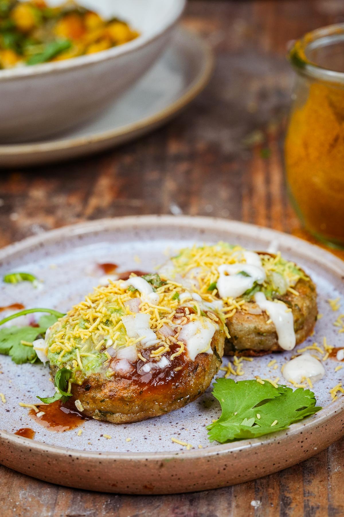 A plate holds two spiced patties garnished with shredded cheese and creamy sauces. A glass jar with a yellow powder and a white bowl of food are in the background on a wooden surface.