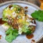 A plate of two aloo tikki chaat pieces topped with yogurt, tamarind chutney, green chutney, sev, chopped onions, and garnished with fresh cilantro leaves.
