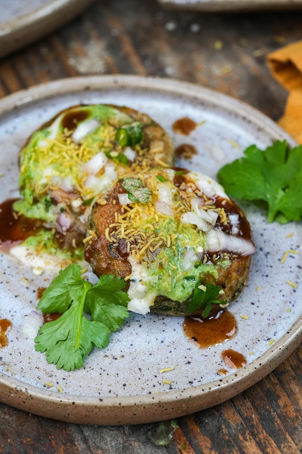 A ceramic plate with two pieces of aloo tiki chaat, garnished with chopped onions, sev, coriander leaves, and various sauces.