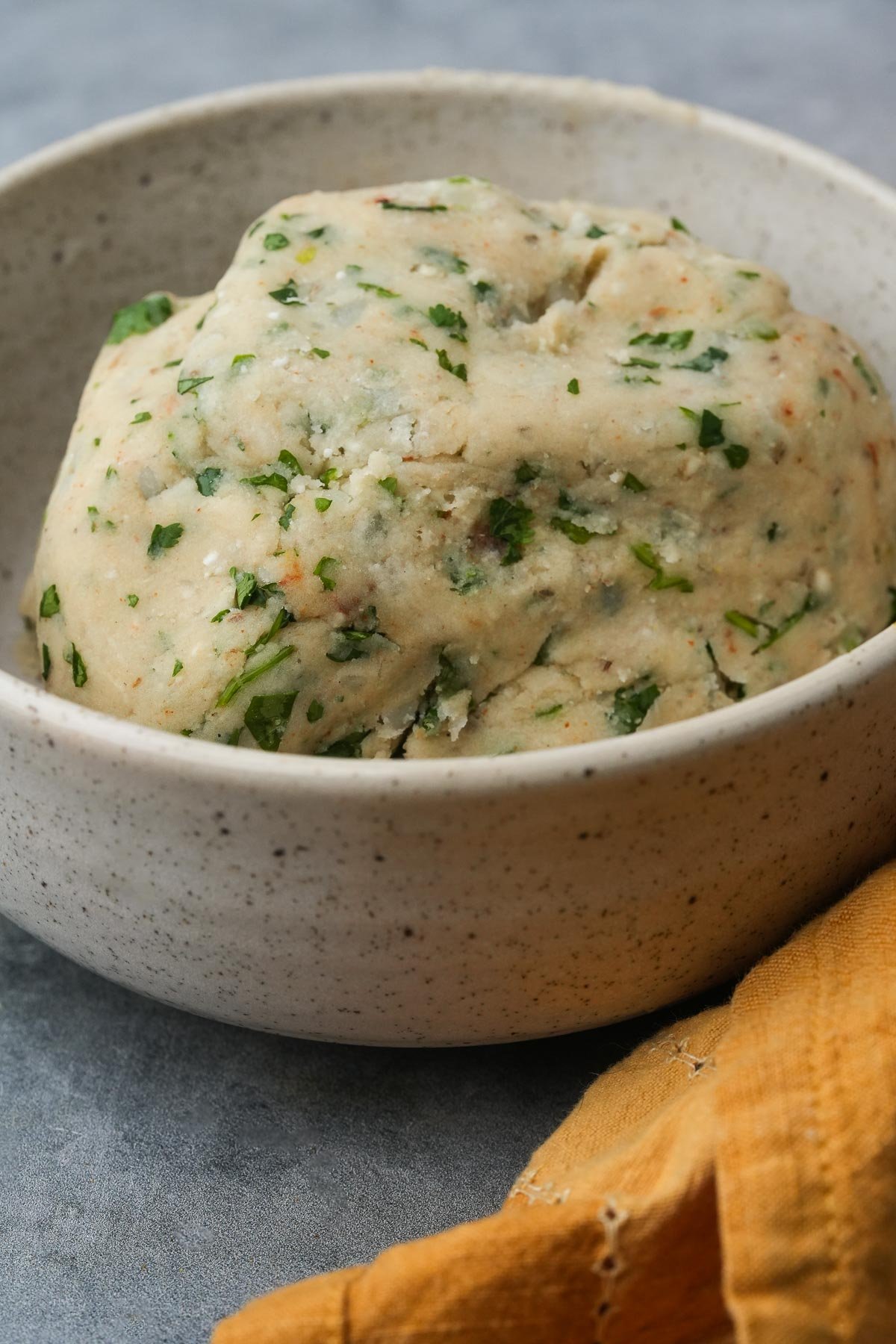 A bowl of dough for making aloo tikki mixed with chopped herbs, placed on a grey surface with a yellow cloth beside it.