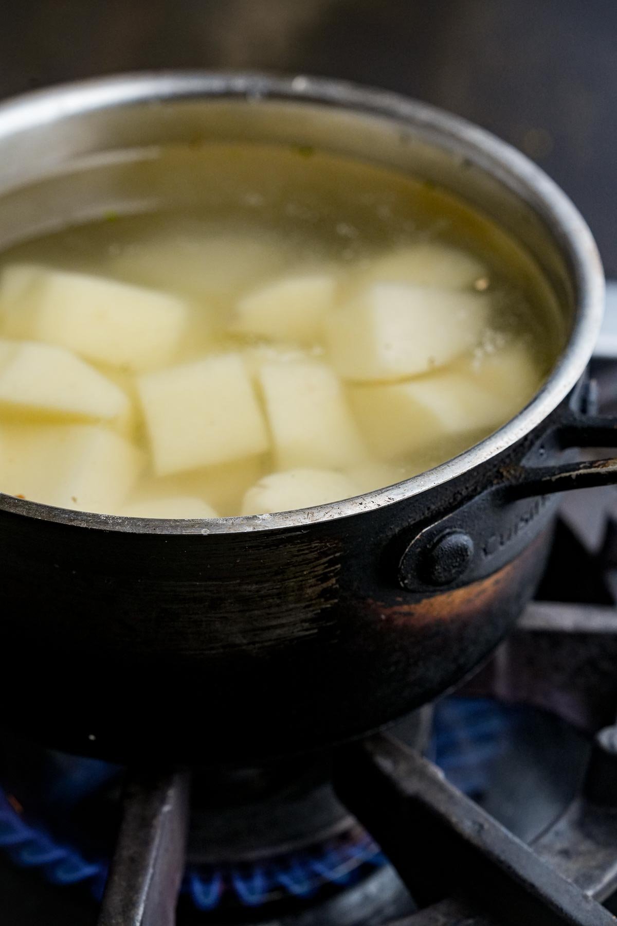 A pot of cubed potatoes boiling in water on a gas stove with a blue flame.