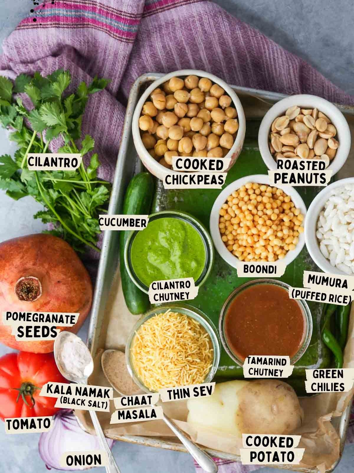 A tray contains ingredients for bhel puri including cooked chickpeas, roasted peanuts, boondi, murmura, two chutneys, thin sev, and garnishes including cilantro, cucumber, pomegranate seeds, tomato, chopped onion, and cooked potato.