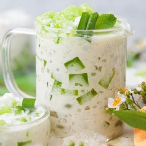 A glass cup filled with creamy Buko Pandan garnished with dicedpandan jelly, alongside small white and yellow flowers.