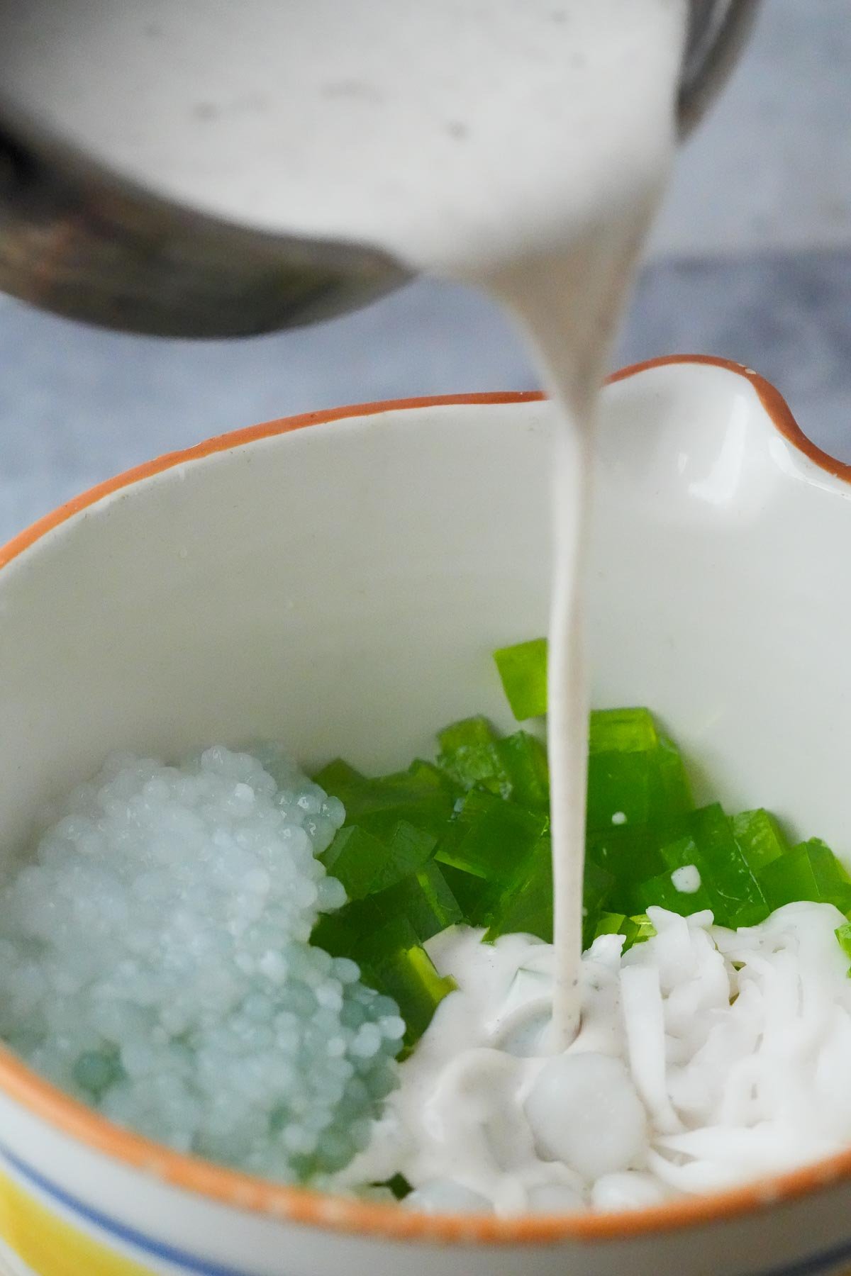 A bowl contains green pandan jelly cubes, white tapioca pearls, palm fruits, and coconut. A creamy white coconut sauce is being poured into the bowl.