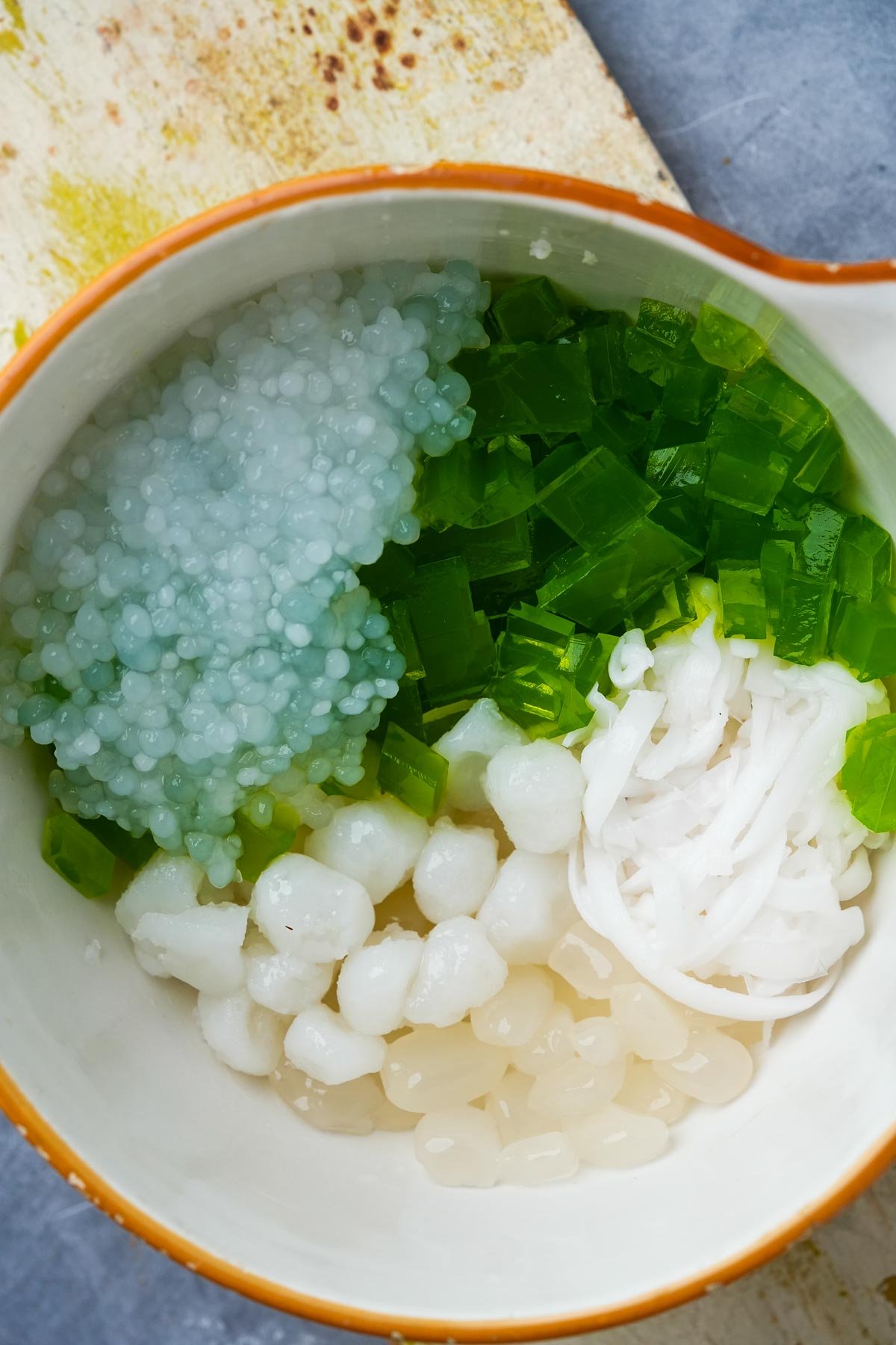 A bowl containing cooked sago pearls, green pandan jelly cubes, palm fruits,, and white shredded young coconut.