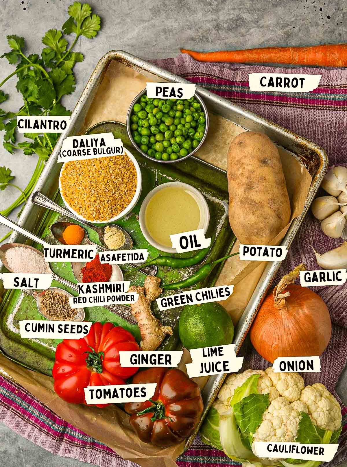 A tray filled with labeled ingredients including cilantro, peas, carrot, potato, oil, turmeric, asafetida, salt, daliya, Kashmiri red chili powder, green chilies, cumin seeds, garlic, lime juice, tomatoes, ginger, onion and cauliflower.