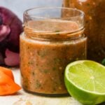 A glass jar filled with homemade habanero salsa, accompanied by a lime slice and a whole tomato, set on a rustic kitchen countertop.