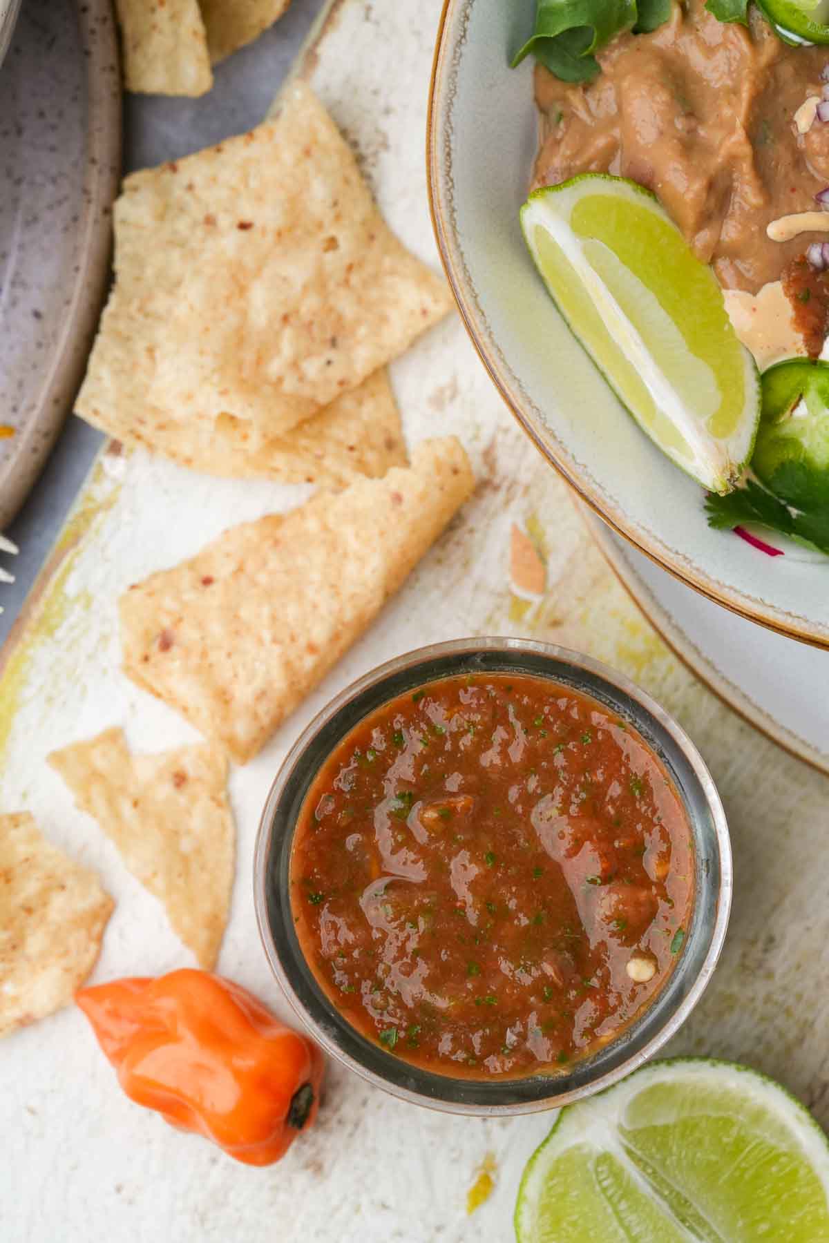 A bowl of habanero salsa with tortilla chips, lime wedges, and a chili pepper on a textured surface.