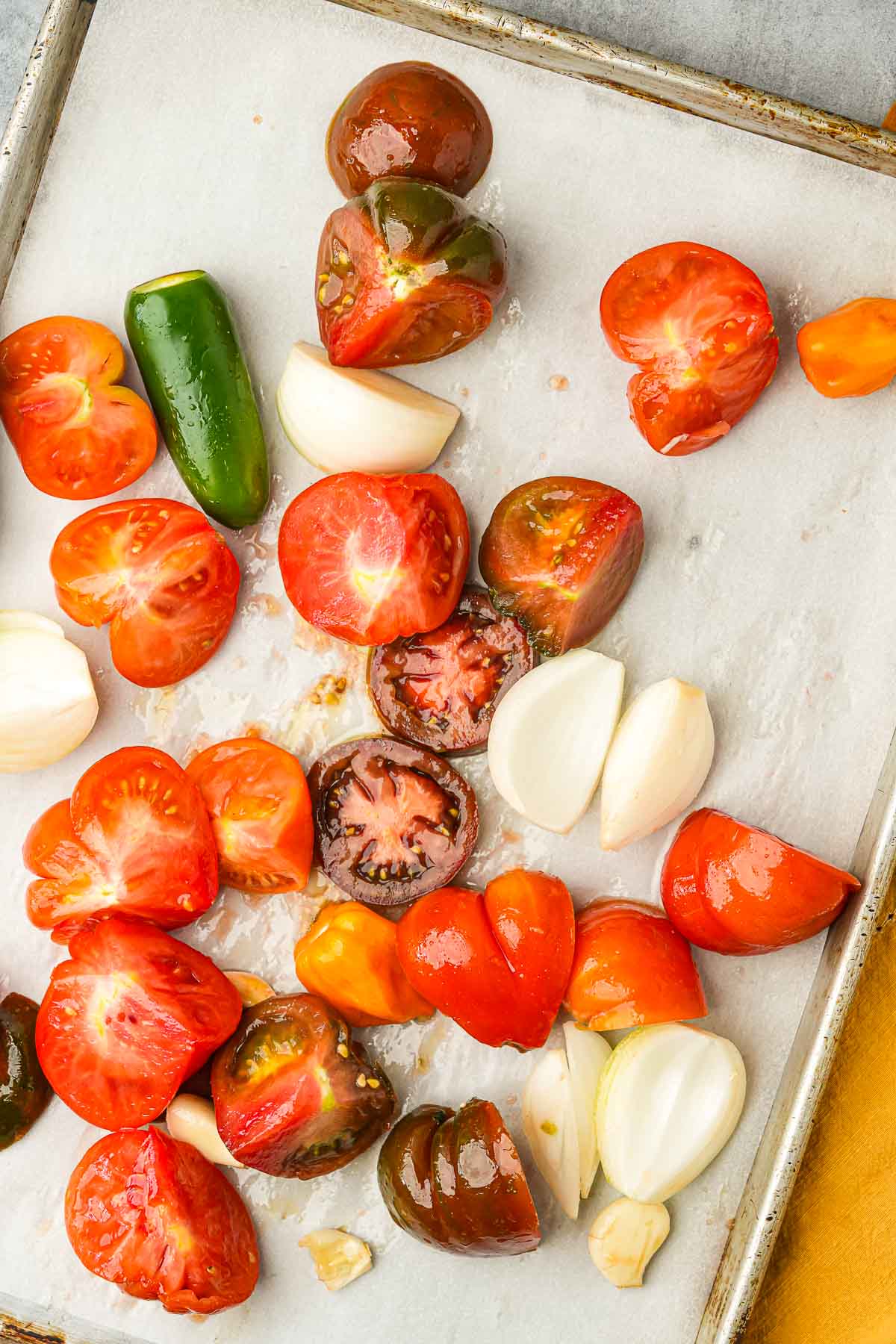 Halved tomatoes, a jalapeno, and garlic cloves arranged on a parchment-lined baking sheet.