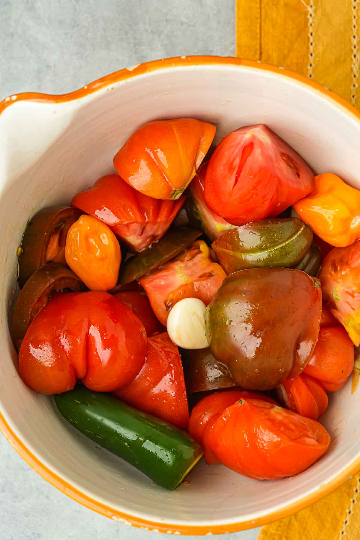 A bowl containing assorted tomatoes and a whole garlic clove, with visible cut marks on the tomatoes, viewed from above.