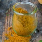 A glass jar filled with yellow Madras Curry Powder, some of which is spilled on the wooden surface.