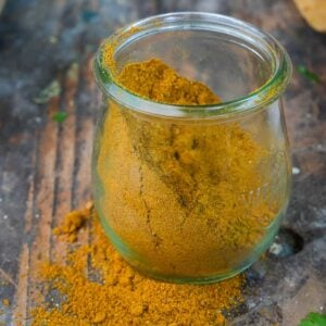 A glass jar filled with yellow Madras Curry Powder, some of which is spilled on the wooden surface.