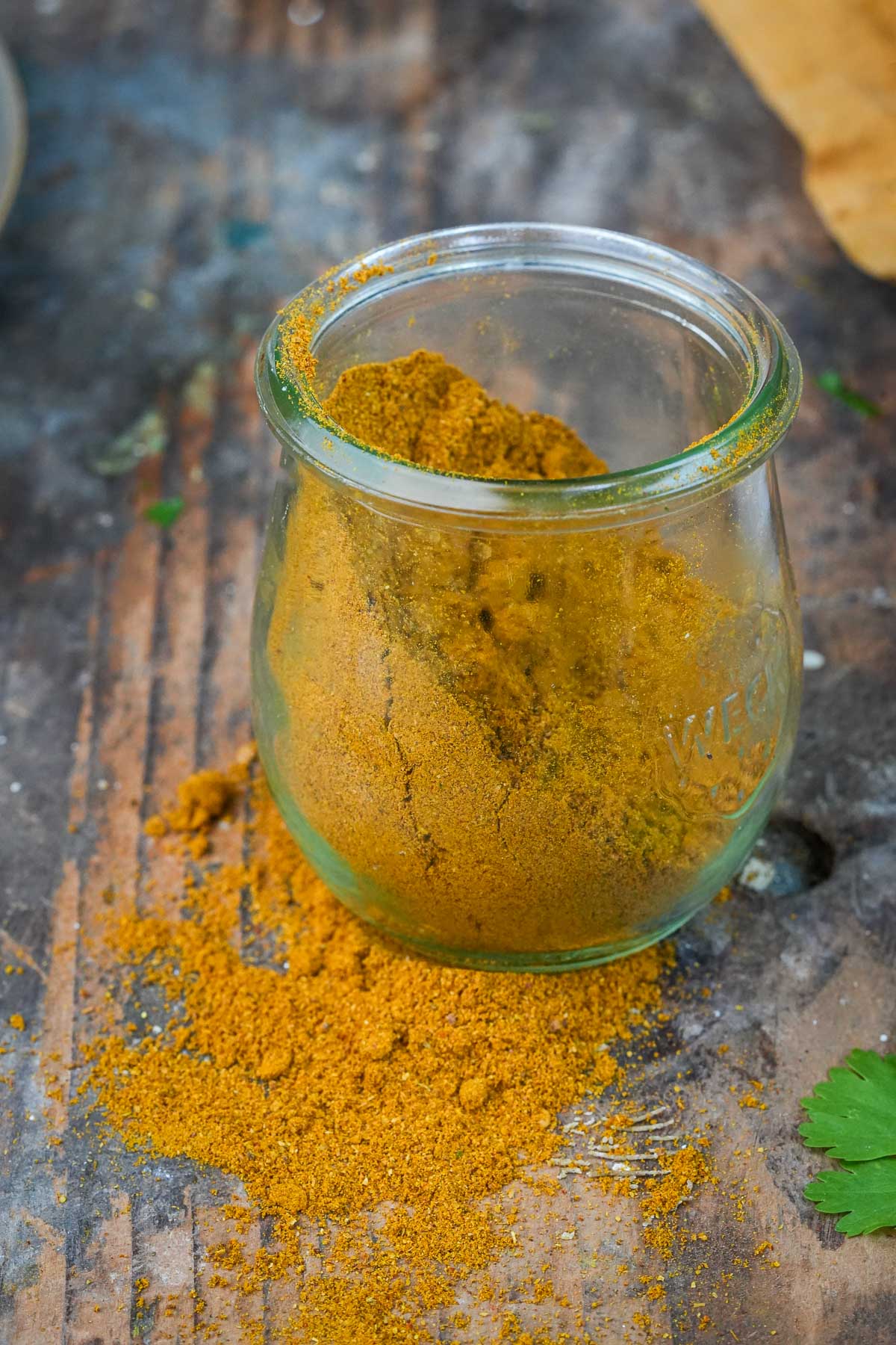 A small glass jar filled with Madras Curry Powder, some of which has spilled onto a wooden surface.