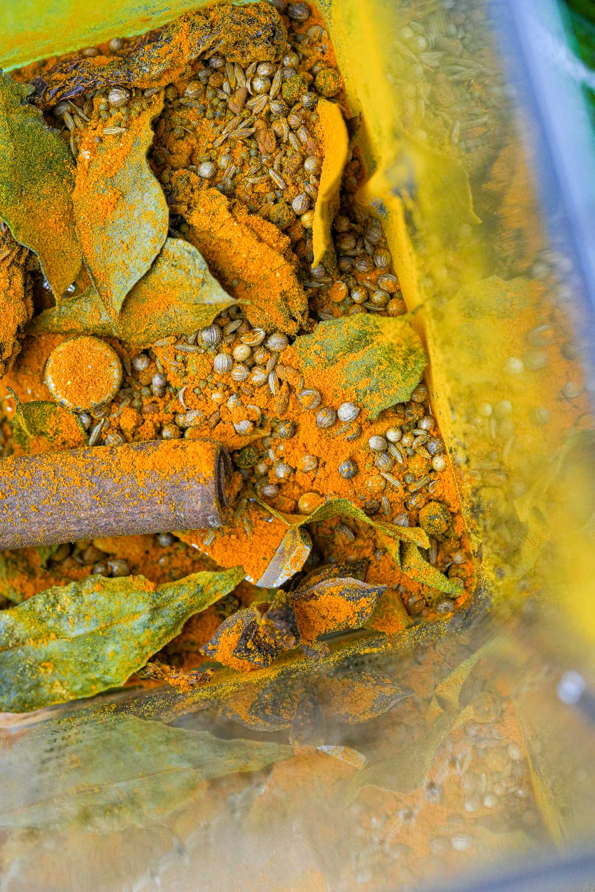 A close-up of a blender filled with spices for making Madras Curry Powder, including bay leaves, turmeric powder, coriander seeds, and a cinnamon stick.