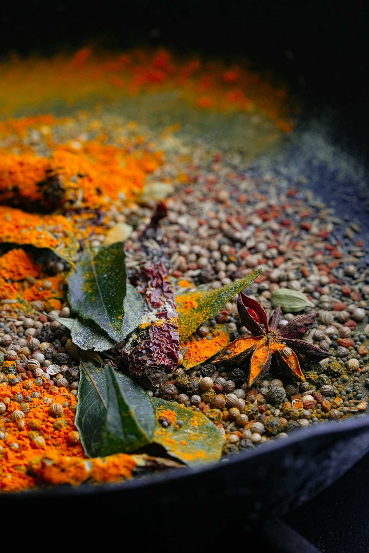 A close-up of various colorful spices, including turmeric, coriander seeds, curry leaves, and dried chili peppers, toasting in a cast iron pan.