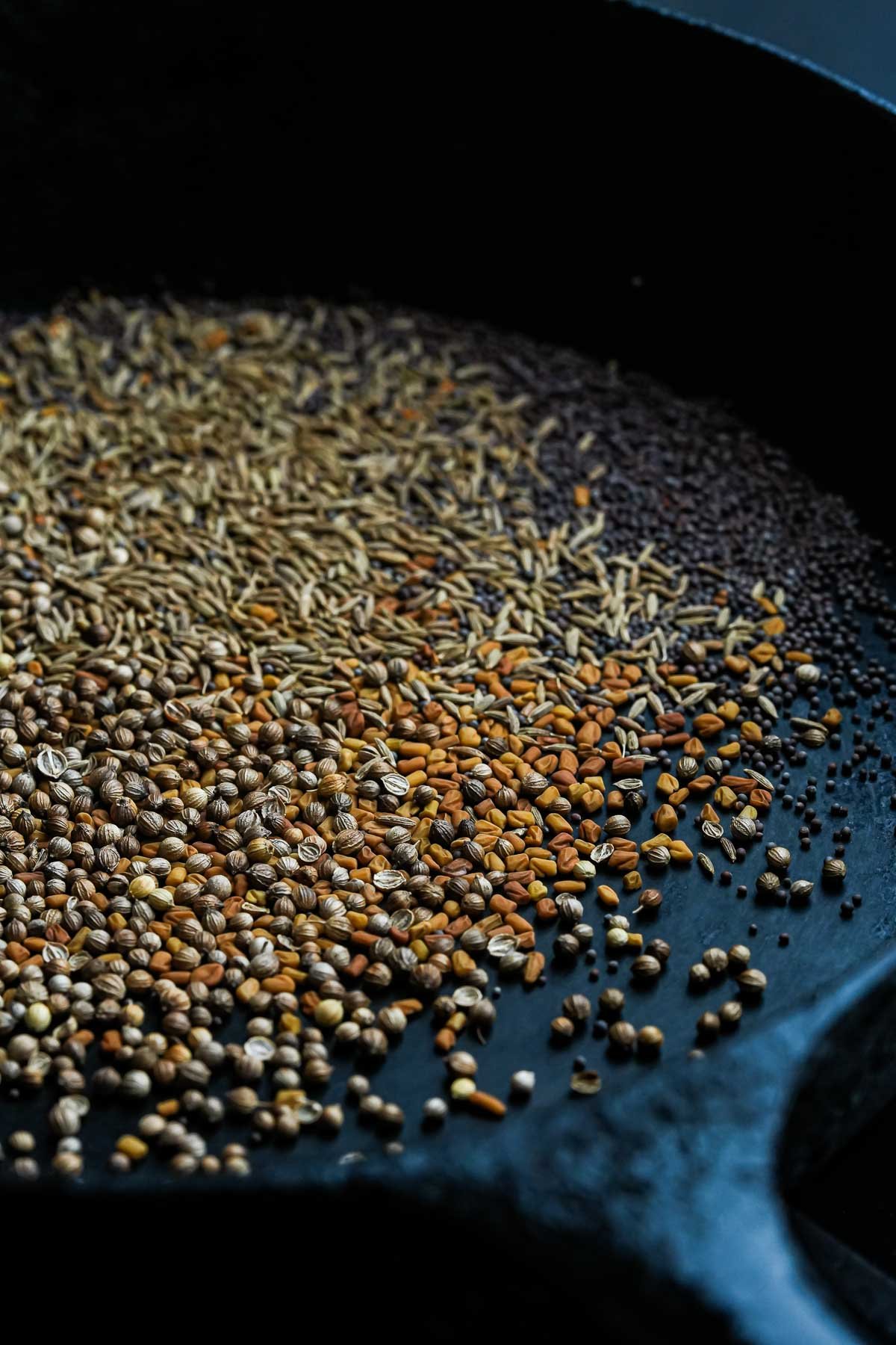 A close-up of a black skillet filled with various whole spices, including coriander seeds, cumin seeds, mustard seeds, and fenugreek seeds.