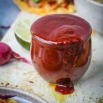 A glass of new mexico style Red Chili Sauce with a lime wedge on the side, displayed on a plate with colorful spices.