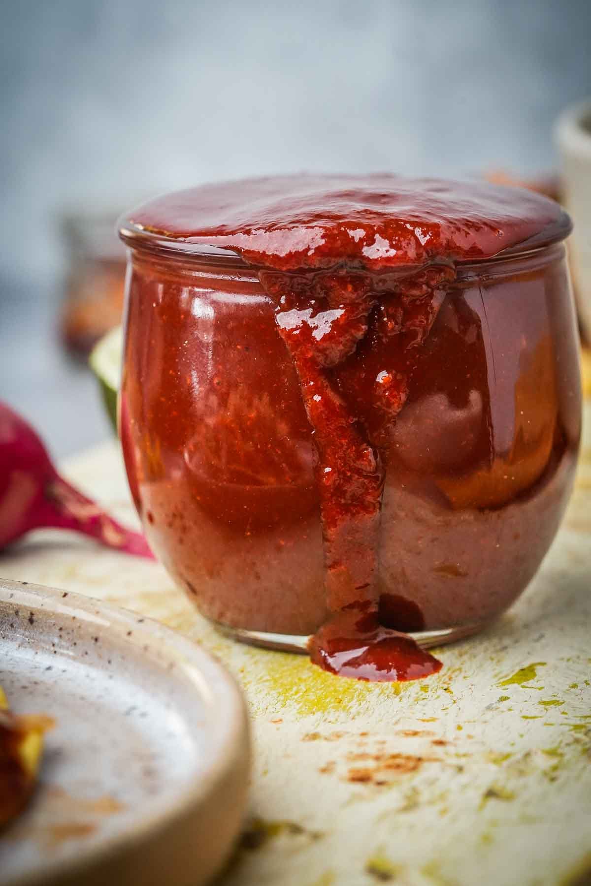A glass jar overflowing with thick Red Chili Sauce on a lightly stained surface, with a smudge of sauce on the jar's edge.