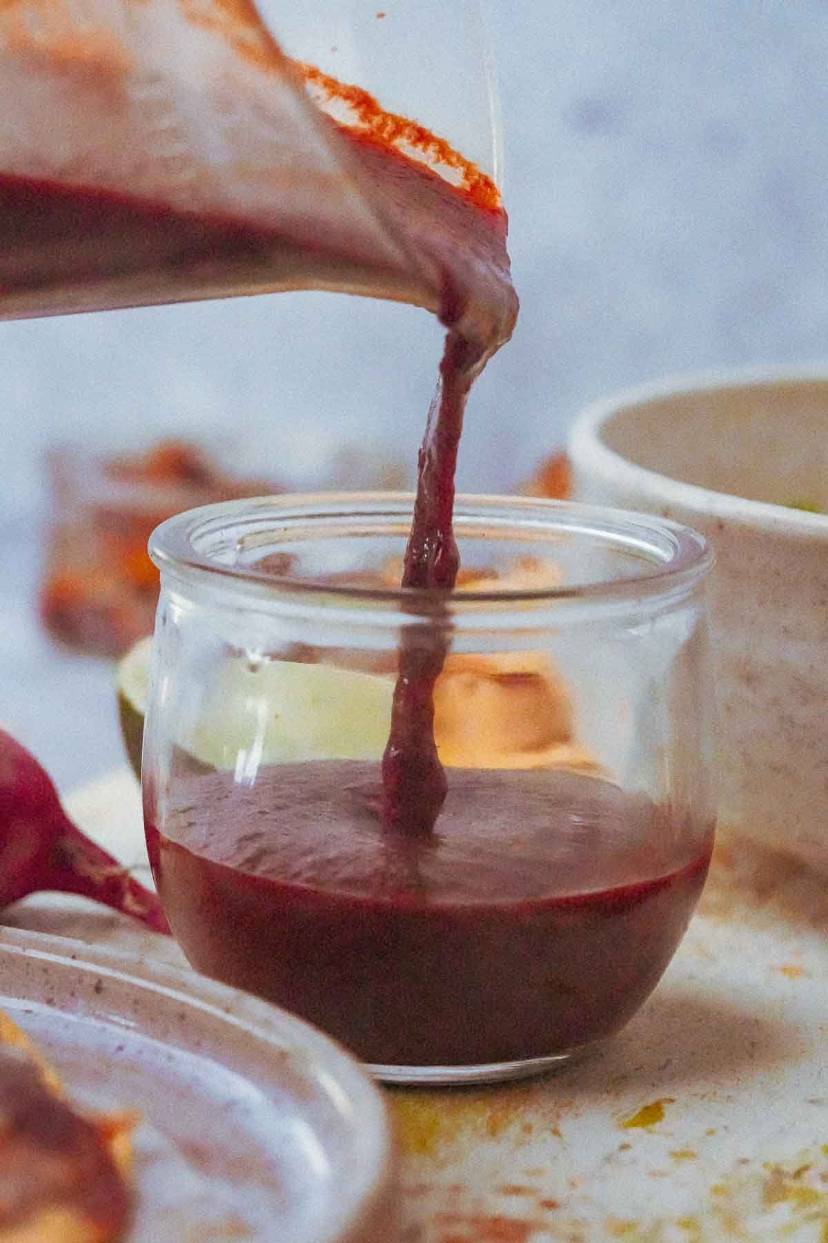 Pouring dark Red Chili Sauce into a small glass jar on a speckled countertop, surrounded by spices and cooking utensils.