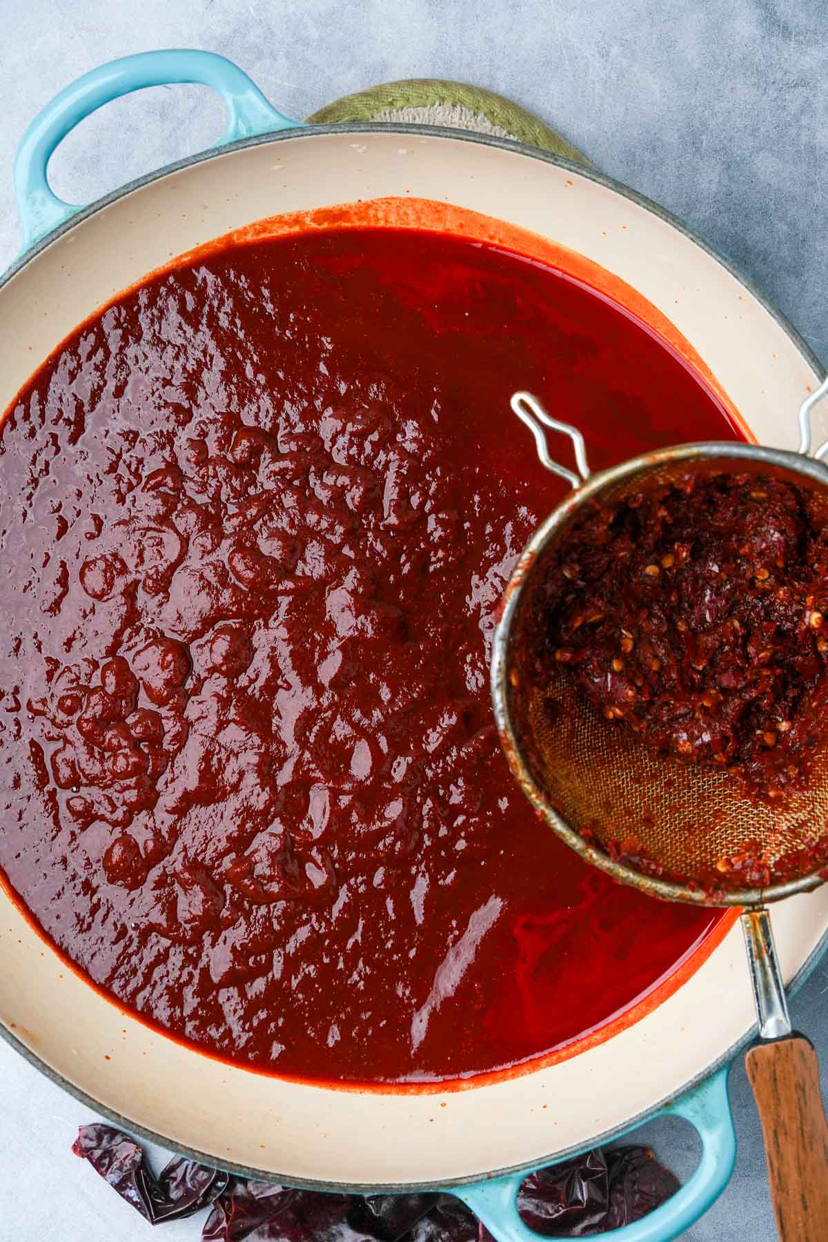 A large pot containing deep red enchilada sauce, with a sieve holding residue over the pot.