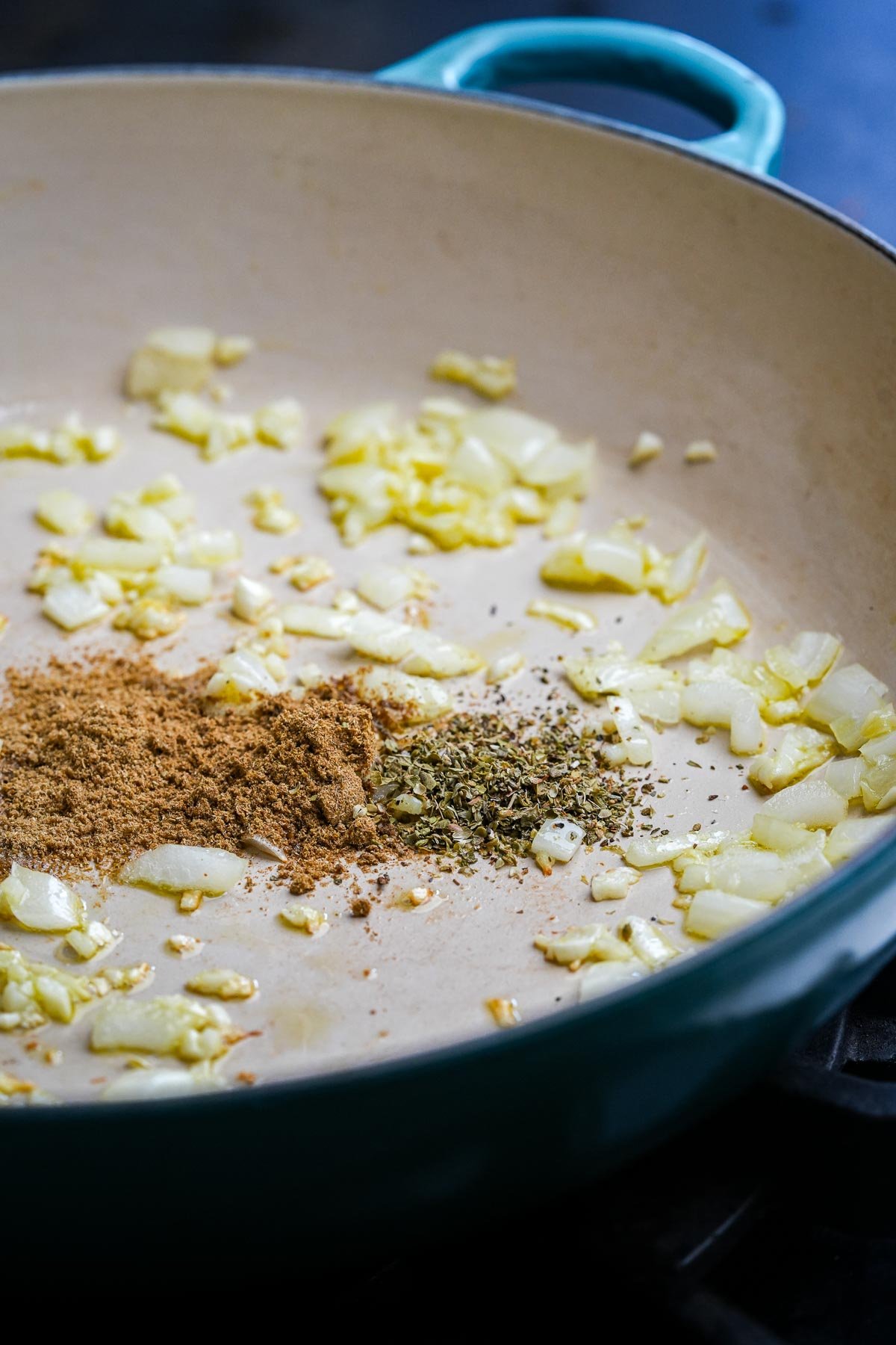 Diced onions, garlic, and spices including cumin and oregano cooking in a light blue enamel pan.