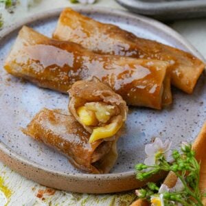 A plate with three caramel-coated Turon, one of which is cut open to reveal a banana filling. Small flowers are visible near the plate.