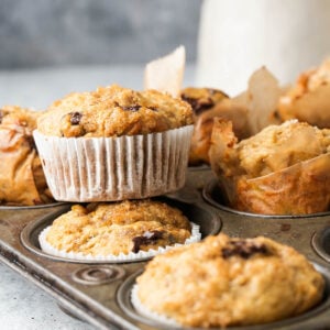 A close-up of freshly baked Vegan Banana Muffins in a muffin tin, with one muffin in a white paper liner, topped with a crumbly texture featuring visible raisins or chocolate chips.