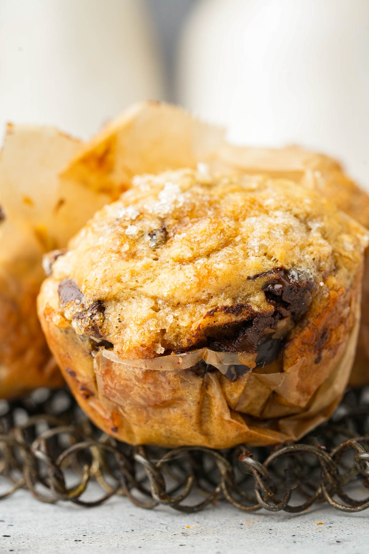 A close-up of a Vegan Banana Muffin with chocolate chunks and walnuts in a paper liner, sprinkled with sugar on top, resting on a wire rack.