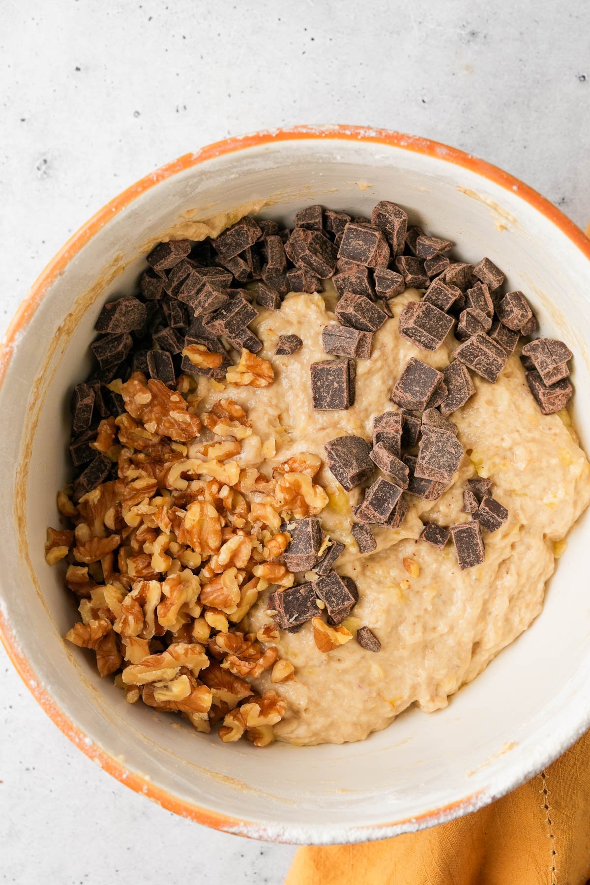 A mixing bowl contains banana muffin batter with chopped walnuts and dark chocolate chunks.