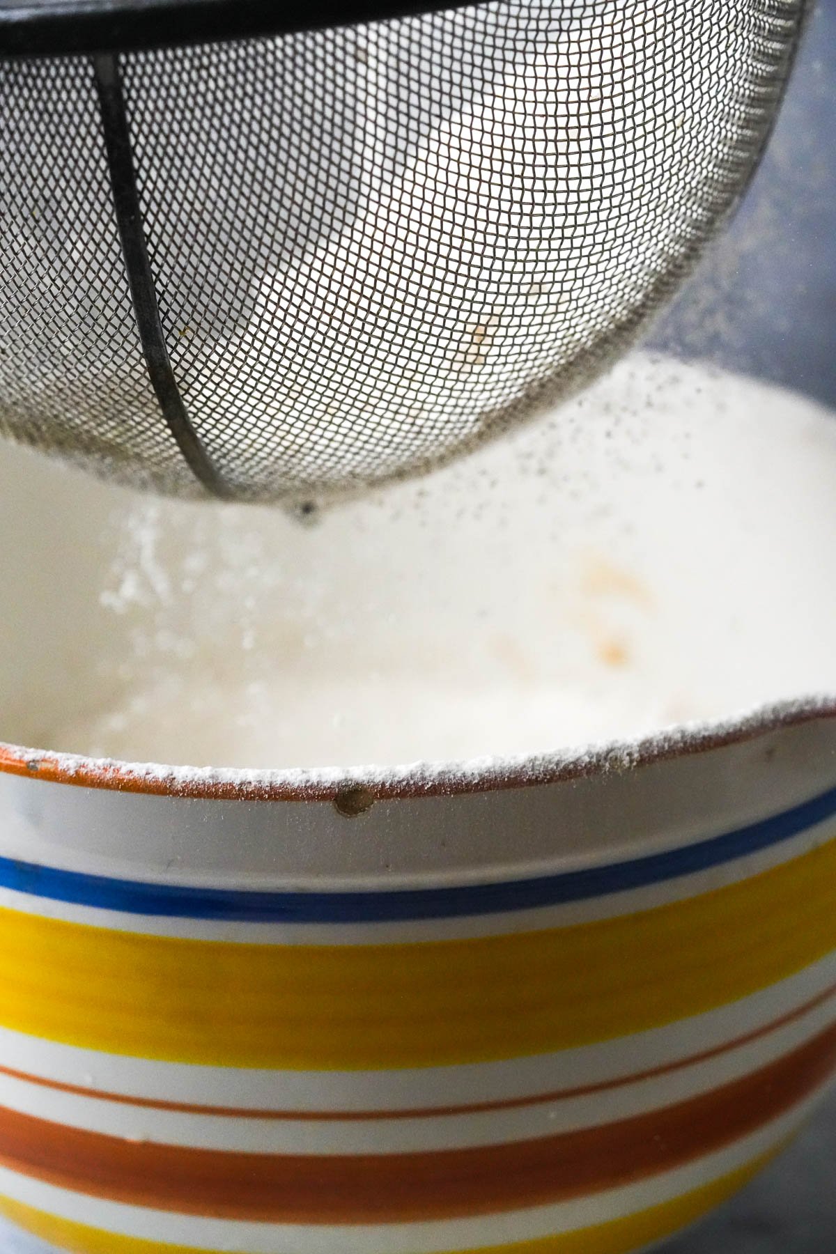 A sifter releasing flour into a multicolored striped bowl.