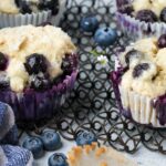 Three vegan blueberry muffins in paper liners are placed on a cooling rack. Fresh blueberries are scattered around the muffins. A cloth napkin is partially visible to the left.