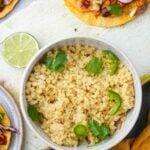 A bowl of Vegan Cotija Cheese garnished with lime slices and cilantro, accompanied by a plate of tacos in the background.