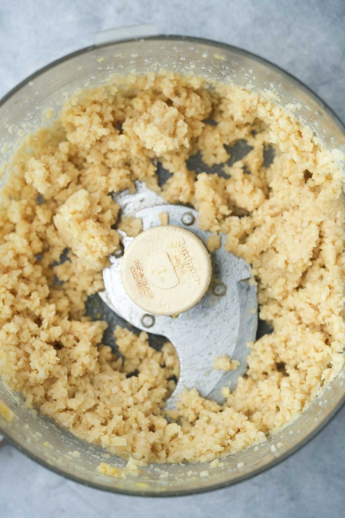 Ground vegan cotija in a food processor, with visible crumbly texture and blades in view.