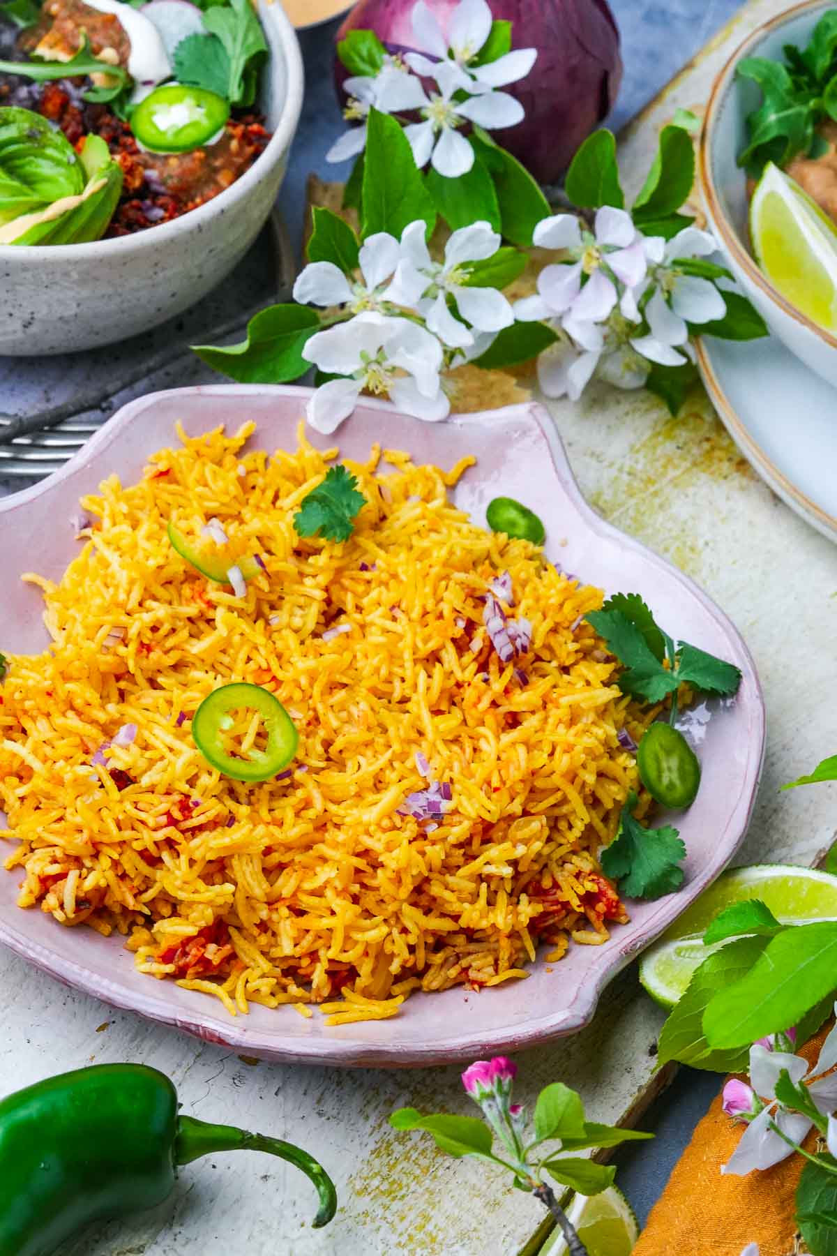A vibrant dish of seasoned Vegan Mexican Rice with sliced jalapeños and herbs, accompanied by other vegan Mexican dishes and floral decorations on a textured wooden table.