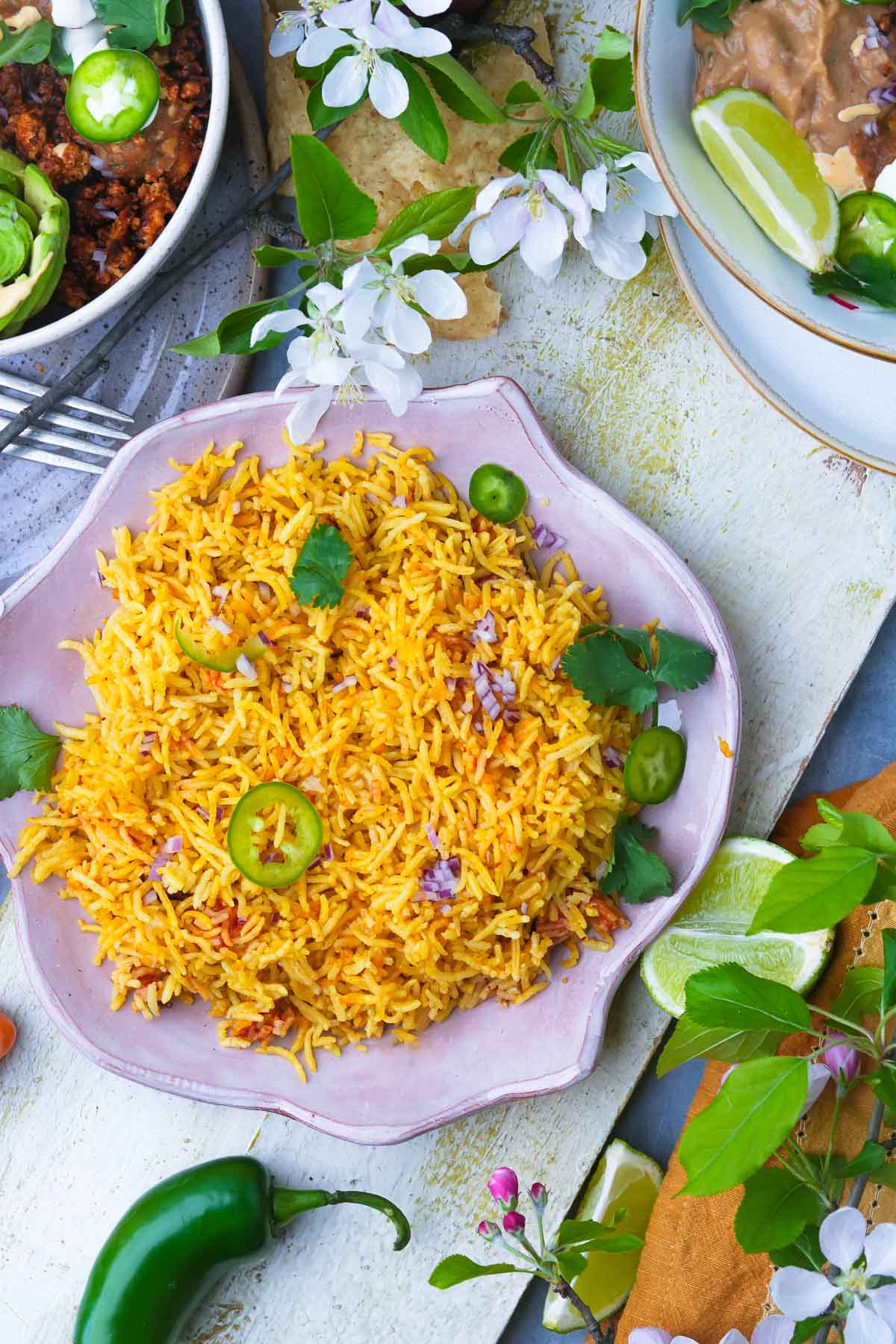 A plate of vibrant yellow Vegan Mexican Rice garnished with green chili slices and herbs, surrounded by assorted dishes and fresh flowers on a decorated table.