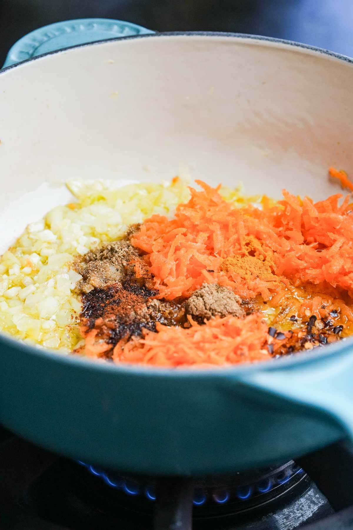 Carrots, onions, and spices sautéing in a light blue enamel pot on a stove.
