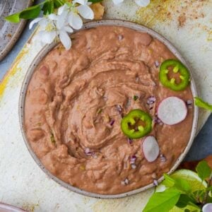 A bowl of creamy vegan refried beans topped with sliced jalapeños and radishes, garnished with flower petals, on a rustic table.