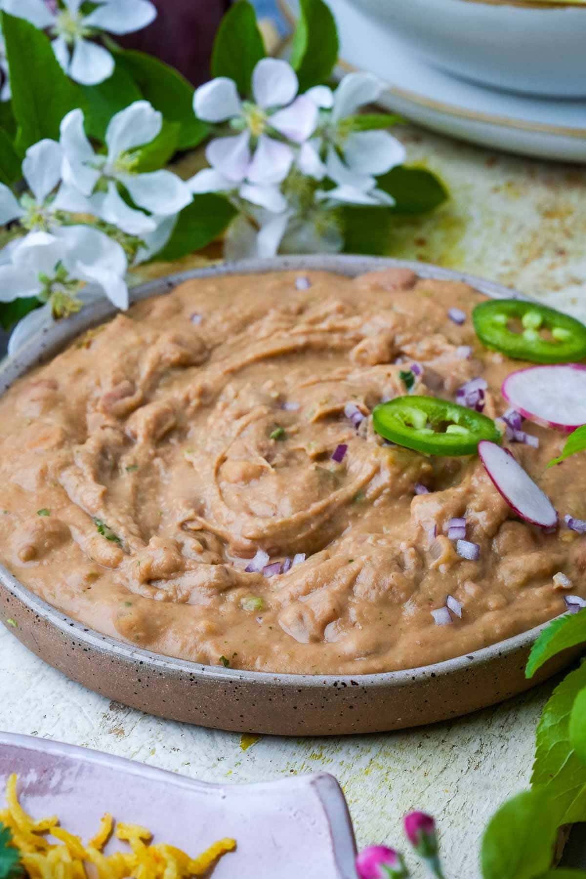 A bowl of creamy vegan refried beans garnished with sliced radishes and onions, accompanied by fresh blossoms on a rustic table.