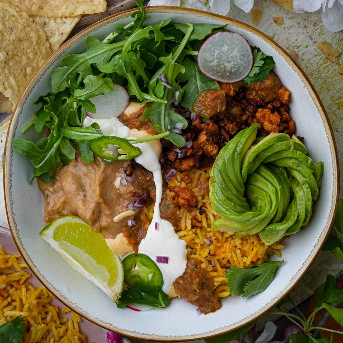 A vegan taco bowl including rice, refried beans, avocado, salad greens, and tofu chorizo, garnished with lime and served with tortilla chips.