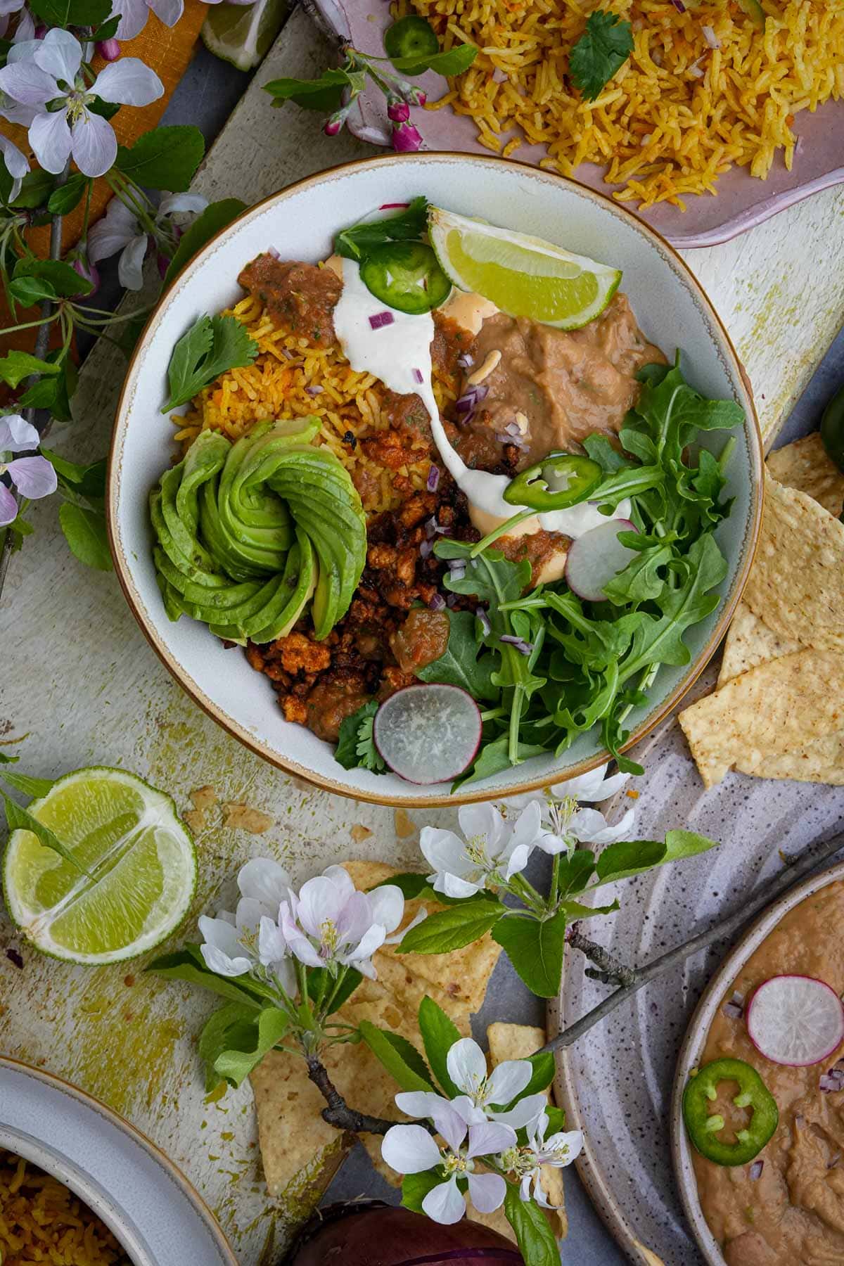 A colorful Mexican vegan taco bowl featuring Mexican rice, vegan taco meat, avocado slices, and salad, garnished with lime and surrounded by flowers.