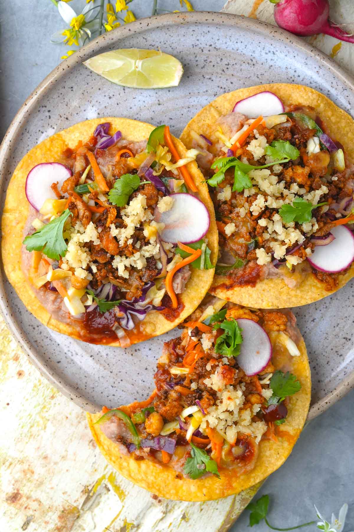 A plate with three Vegan Tostadas topped with plant-based chorizo crumbles, beans, cabbage slaw, almond cotija cheese, and slices of radish, garnished with cilantro.