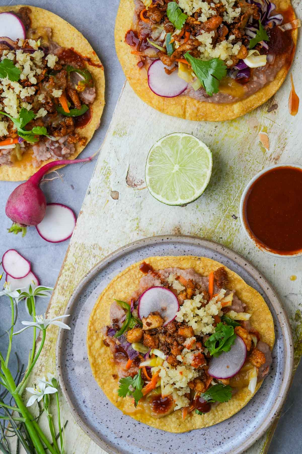 A ramekin of red chili sauce next to three vegan Tostadas topped with tofu chorizo, beans, vegan cotija cheese, and topped with radish slices on a circular ceramic plate.