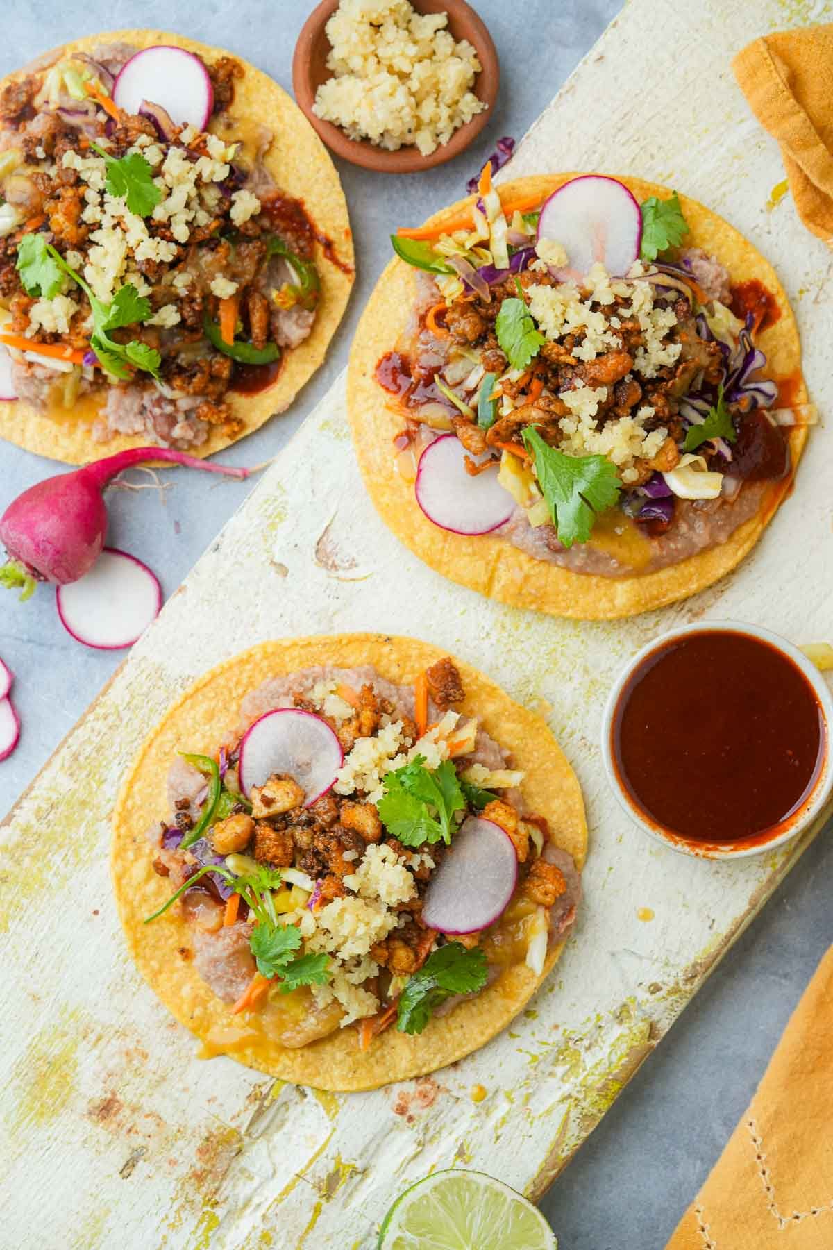 Three fully assembled Vegan Tostadas topped with plant-based chorizo crumbles, beans, cabbage slaw, almond cotija cheese, and slices of radish, garnished with cilantro.